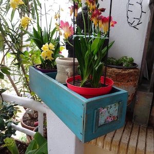 May include: A turquoise wooden planter box with a distressed finish, holding a red pot with green plants and colorful flowers. The box has a decorative floral panel and is suspended from a metal frame. Other potted plants are visible in the background.