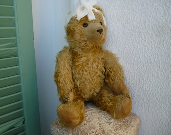 Puede incluir: Un oso de peluche de color marrón dorado con pelaje largo y desgreñado, ojos oscuros y nariz cosida. El oso presenta almohadillas de tela marrón claro en las patas y una venda de gasa blanca alrededor de la cabeza, atada con un nudo.