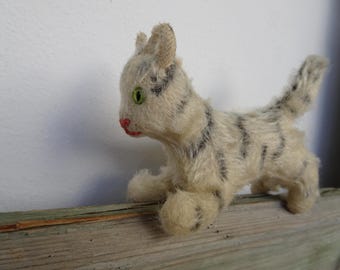May include: A vintage stuffed cat toy with a cream-colored body and black stripes. The cat has green eyes and a red nose. It is posed on a wooden surface, with a white wall in the background.