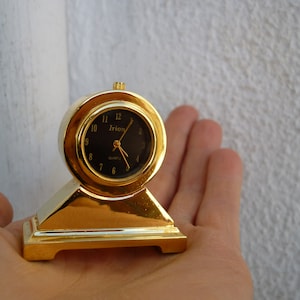 May include: A gold-toned desk clock with a black face and white hands. The clock reads "Irion Quartz".