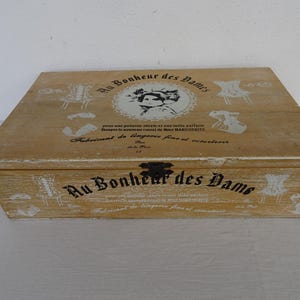 Puede incluir: Una caja rectangular de madera con acabado marrón claro, con gráficos impresos en blanco y negro de corsés y un retrato. El texto dice Au Bonheur des Dames, Fabricant de lingerie fine et corseterie y Mme MARGUERITE. Tiene un cierre de metal.
