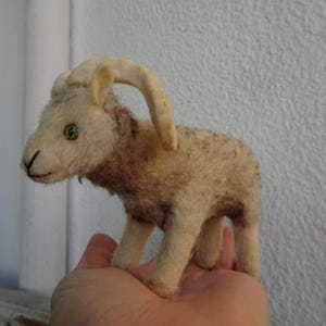 May include: A white and brown plush toy goat with a curled horn. The goat has green eyes and is standing on a hand.