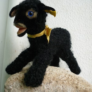 Könnte beinhalten: Ein Vintage-Spielzeug in Form eines schwarzen Lamms mit blauen Augen und einem gelben Band um den Hals. Das Lamm mit seinem lockigen Fell steht auf einer Steinfläche. Der Mund des Spielzeugs ist geöffnet.