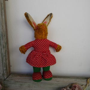 Può includere: Un giocattolo vintage a forma di coniglio di peluche, visto da dietro, con un vestito rosso a pois bianchi. Il coniglio ha la pelliccia marrone, orecchie lunghe, leggings verdi e scarpe rosse a pois. Il giocattolo è posizionato su una superficie di legno.