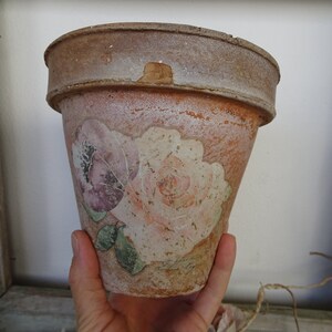 May include: A distressed terracotta flower pot with a faded floral design of pink and purple roses. The pot has a weathered appearance, adding to its vintage charm. Suitable for planting flowers or herbs.