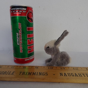 Puede incluir: Una lata de bebida energética Hell Strong Watermelon junto a una pequeña figura de conejo gris y blanco. Una regla de madera con el texto "AUTOMOBILE TRIMMINGS NAUGAHYDE" está en primer plano.