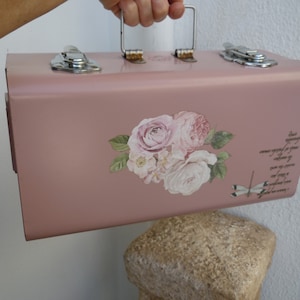 Puede incluir: Una caja de herramientas de metal rosa con asa y cierres plateados. La caja de herramientas está decorada con una calcomanía floral con rosas rosas y blancas y hojas verdes. También se ve una libélula y texto.