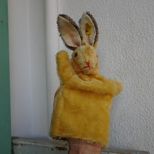 Könnte beinhalten: Eine Vintage-Handpuppe in Form eines gelben Kaninchens mit langen Ohren und einer rot genähten Nase. Die Puppe hat eine weiche, plüschige Textur und ist für die Hand konzipiert, zum Spielen oder zur Dekoration.