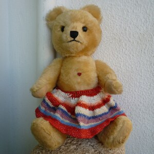 Peut inclure: Un ours en peluche vintage avec une fourrure brun clair, des yeux et un nez noirs, et un petit cœur rouge sur la poitrine. L'ours porte une jupe en crochet rayée colorée rouge, blanche, bleue et violette. L'ours est assis sur une surface texturée.