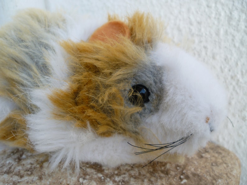 May include: A plush toy of a guinea pig with white, brown, and gray fur. The toy has black whiskers and a small, pink nose.