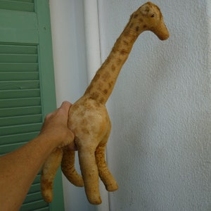 Può includere: Un giocattolo di peluche di giraffa di colore beige con macchie marroni. La girafa è in piedi su una superficie bianca e ha un collo e gambe lunghe.