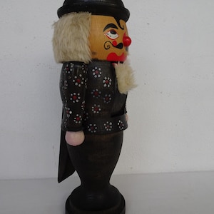 May include: A wooden nutcracker figurine featuring a black bowler hat, a painted face with a red nose and mustache, and a black jacket adorned with small white and red floral patterns. It has synthetic hair and a dark, pedestal-style base.