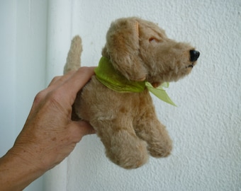 Puede incluir: Un perro de peluche marrón claro con una cinta verde alrededor del cuello. El perro está sentado sobre una superficie blanca.