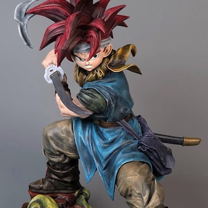 May include: A detailed figurine of a warrior in a dynamic pose, wielding a sword. The figure has spiky red hair, a blue tunic, and brown pants. The base features a rocky terrain with green and yellow accents. The overall aesthetic is reminiscent of fantasy art.