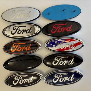 May include: Ten oval-shaped Ford emblems in various designs, including chrome, black, red, and American flag patterns. Each emblem features the Ford script logo. One emblem is shown from the back, revealing a blue adhesive backing and three pins.
