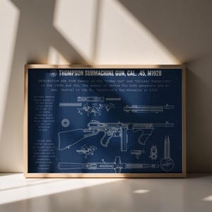 May include: A framed blueprint-style poster featuring a white line drawing of a Thompson Submachine Gun, Cal. .45, M1928 on a dark blue background. The print includes technical diagrams, component details, and historical text about the firearm.