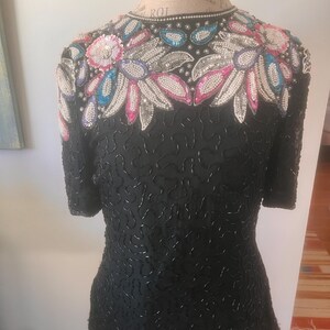 May include: Black short-sleeved top with intricate beadwork. The neckline and shoulders are adorned with floral designs in pink, blue, and silver sequins and beads. The body of the top is covered in small, black beads.