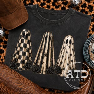 Könnte beinhalten: Ein anthrazitfarbenes T-Shirt mit drei Geisterfiguren in schwarz-beigen Mustern, darunter Karos, Streifen und Sterne. Jeder Geist ist an der Basis mit schwarzen Rosen verziert. Der Text ATD DESIGNS ist unten rechts in Weiß aufgedruckt.