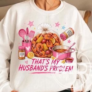 May include: White sweatshirt featuring a seafood boil graphic with shrimp, corn, and seasoning. Pink text reads "THAT'S MY HUSBAND'S PROBLEM!" with pink stars and cooking utensils. A fun, food-themed design.