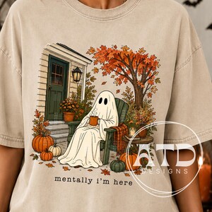 Puede incluir: Una camiseta beige con un gráfico de un fantasma blanco sentado en una silla en un porche, sosteniendo una taza. La escena incluye calabazas naranjas, hojas de otoño, un árbol y una puerta. El texto "mentally i'm here" está impreso debajo del gráfico.