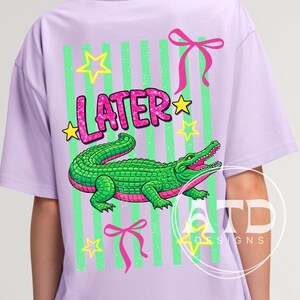 Può includere: T-shirt lilla con un design di alligatore verde, dettagli rosa e la scritta "LATER" in glitter rosa. Il design include stelle gialle, strisce verticali verdi e fiocchi rosa. Abbigliamento casual.