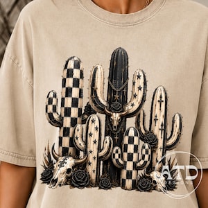 K&ouml;nnte beinhalten: Ein beiges T-Shirt mit einem Grafikdruck aus mehreren Kakteen in Schwarz und Creme. Das Design umfasst Karomuster, Sternmotive und drei Tiersch&auml;del, die mit Ketten und schwarzen Rosen verziert sind, mit dem Text ATD DESIGNS in der Ecke.