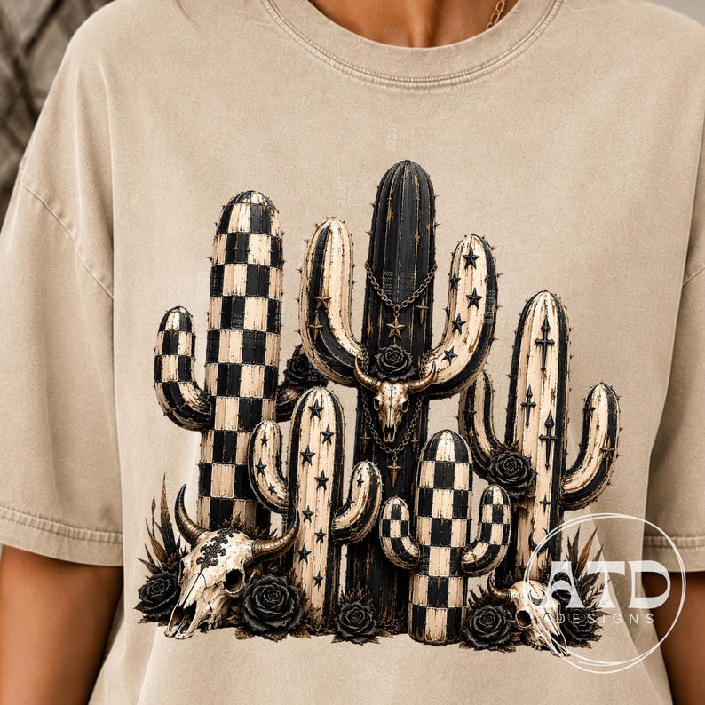 K&ouml;nnte beinhalten: Ein beiges T-Shirt mit einem Grafikdruck aus mehreren Kakteen in Schwarz und Creme. Das Design umfasst Karomuster, Sternmotive und drei Tiersch&auml;del, die mit Ketten und schwarzen Rosen verziert sind, mit dem Text ATD DESIGNS in der Ecke.