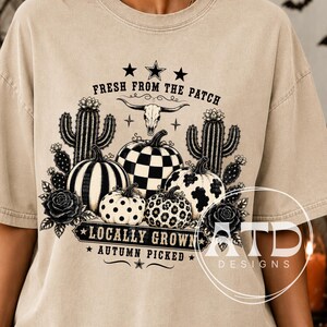 May include: A beige t-shirt featuring a black graphic print of pumpkins, cacti, roses, and a steer skull. The text reads FRESH FROM THE PATCH, LOCALLY GROWN, and AUTUMN PICKED. The pumpkins display various patterns including checkers, spots, and leopard print.