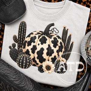 Könnte beinhalten: Ein hellgraues T-Shirt mit einer zentralen Grafik eines cremefarbenen Kürbisses mit schwarzen Kuhflecken, umgeben von mehreren schwarzen Kakteen und zwei hellen Blumen. Die Grafik enthält den Text ATD DESIGNS in einem Kreis.