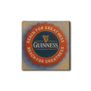 Op de afbeelding: Vierkant schilderij van een Guinness-flesdop. De dop is rood met de woorden "REACH FOR GREATNESS" in het wit. Het midden is donkerblauw met het Guinness-logo en een gouden harp. De achtergrond is lichtbeige.