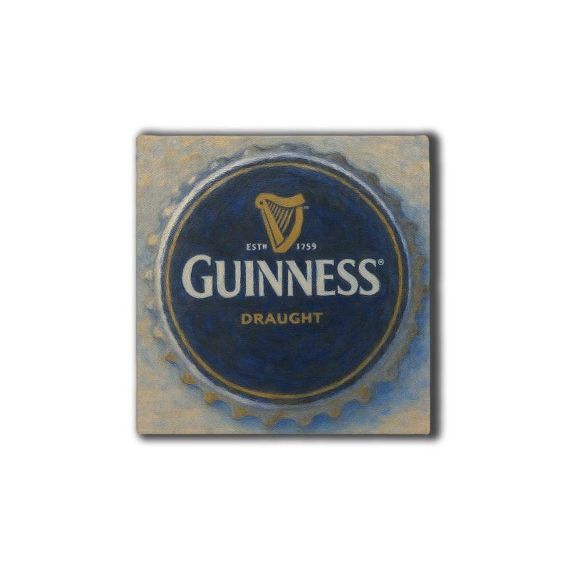 Op de afbeelding: Vierkant kunstwerk met een Guinness Draught flesdop. De dop heeft een donkerblauwe kern met het Guinness logo en een gouden harp. De buitenrand is goudkleurig, tegen een getextureerde, lichtbeige achtergrond.
