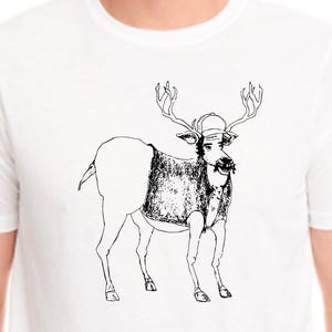 May include: White t-shirt featuring a whimsical black and white illustration of a deer wearing a baseball cap and a sweater. The deer has large antlers and a mustache, creating a humorous design.