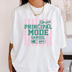 May include: White t-shirt with the text "PRINCIPAL MODE" in green block letters, with a green bow and pink vertical stripes. Below the text is "SAMUEL" and an "OFF" switch graphic.
