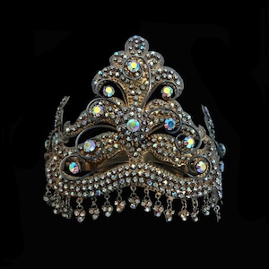 May include: An antique-style crown with a gold-toned metal frame. The crown is adorned with iridescent rhinestones and dangling crystal accents. The background is black.
