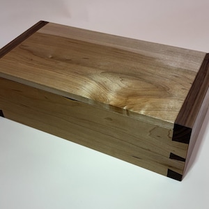 May include: A rectangular wooden box with a light-coloured top and sides, and dark brown accents on the corners. The box features dovetail joints and a natural wood grain finish. The box is made by NJM Woodworks LLC.