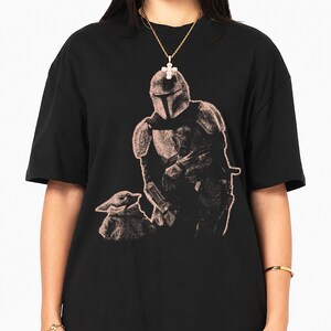 May include: A black short-sleeved t-shirt featuring a light-colored graphic print of a helmeted armored character standing next to a small creature with large ears. The design is rendered in a stippled, textured style on the front of the shirt.