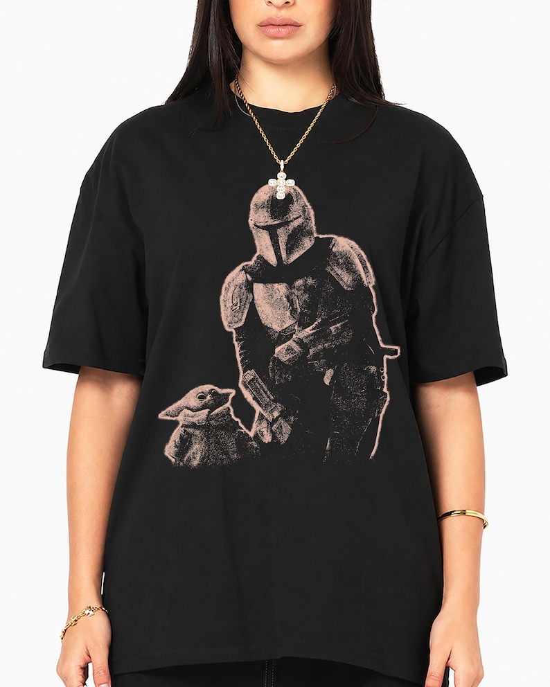 May include: A black short-sleeved t-shirt featuring a light-colored graphic print of a helmeted armored character standing next to a small creature with large ears. The design is rendered in a stippled, textured style on the front of the shirt.