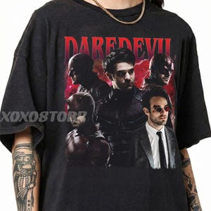 May include: Dark grey t-shirt with a graphic of the Daredevil character in different poses. The word "DAREDEVIL" is printed in red at the top. The brand name "XOXOSTORE" is printed on the left side.