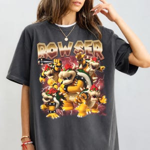 Puede incluir: Una camiseta de manga corta gris carbón con un estampado gráfico del personaje Bowser en varias poses. La palabra BOWSER está impresa en letras grandes, negritas y tonos metálicos sobre el personaje, con un fondo de rayos rojo oscuro.