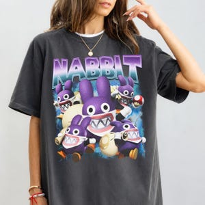 May include: A charcoal gray short-sleeve t-shirt featuring a graphic print of multiple purple rabbit-like characters with large white teeth and wide eyes. The text NABBIT is printed in a purple and white gradient font above the characters.