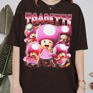 Puede incluir: Una camiseta de manga corta de color marrón oscuro con un estampado gráfico de cinco personajes Toadette de la serie Super Mario. El texto TOADETTE aparece en una fuente llamativa de estilo metálico sobre un fondo de color rojo oscuro.