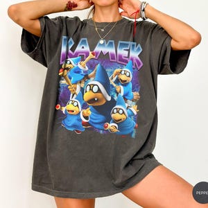 May include: A charcoal grey short-sleeve t-shirt featuring a graphic print of five blue-robed Kamek characters. The text KAMEK is printed in a bold, purple-outlined font above the characters, set against a background of purple lightning bolts.
