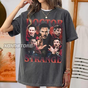 May include: Dark gray t-shirt featuring a graphic of Doctor Strange with the text "DOCTOR STRANGE" in red and orange. The shirt has a vintage, distressed look. The brand name "XOXOSTORE" is printed on the left side of the shirt.
