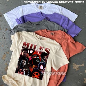 K&ouml;nnte beinhalten: Ein Stapel von f&uuml;nf kurz&auml;rmeligen T-Shirts in Wei&szlig;, Lila, Grau, Orange und Creme. Das cremefarbene Shirt zeigt eine Grafik eines Comic-Charakters mit dem Wort "MILES" in Rot. Der Text "REMEMBER TO CHOOSE COMFORT TSHIRT" steht oben.