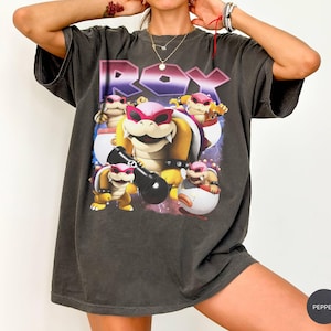 Può includere: Una t-shirt a maniche corte grigio antracite con una stampa grafica del personaggio Roy della serie Super Mario. Il design include il testo ROY in un carattere audace in stile metallico sopra diverse rappresentazioni del personaggio con occhiali rosa.