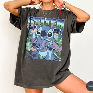 May include: A charcoal gray short-sleeve t-shirt featuring a graphic print of the character Stitch in various poses. The word STITCH is printed in large blue and white letters at the top, set against a background of green lightning bolts.