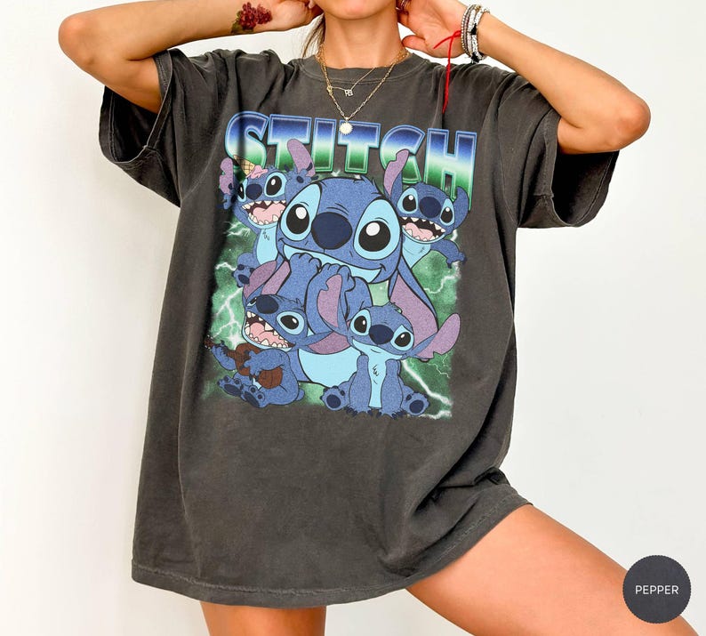 May include: A charcoal gray short-sleeve t-shirt featuring a graphic print of the character Stitch in various poses. The word STITCH is printed in large blue and white letters at the top, set against a background of green lightning bolts.