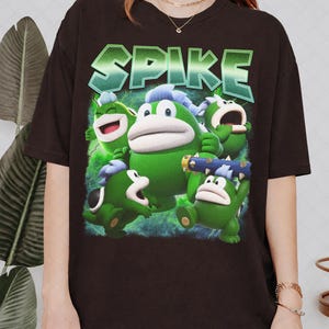 May include: A black short-sleeve t-shirt featuring a graphic print of five green Spike characters from the Super Mario series. The word SPIKE is printed in large, bold, green gradient letters above the characters, set against a green background.