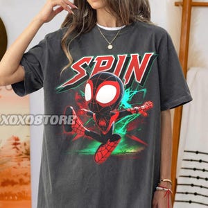 Peut inclure: T-shirt gris foncé avec un graphisme de Spider-Man de dessin animé et le mot "SPIN" en rouge et blanc. Le personnage est dans une pose dynamique avec des détails rouges et noirs, sur un fond turquoise et vert. Le nom de la marque "XOXOSTORE" est imprimé sur le côté gauche.