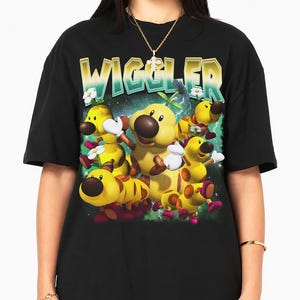 May include: A black short-sleeve t-shirt featuring a graphic print of multiple yellow Wiggler characters from the Super Mario series. The text WIGGLER appears in a large, stylised, gradient font above the characters against a green background.